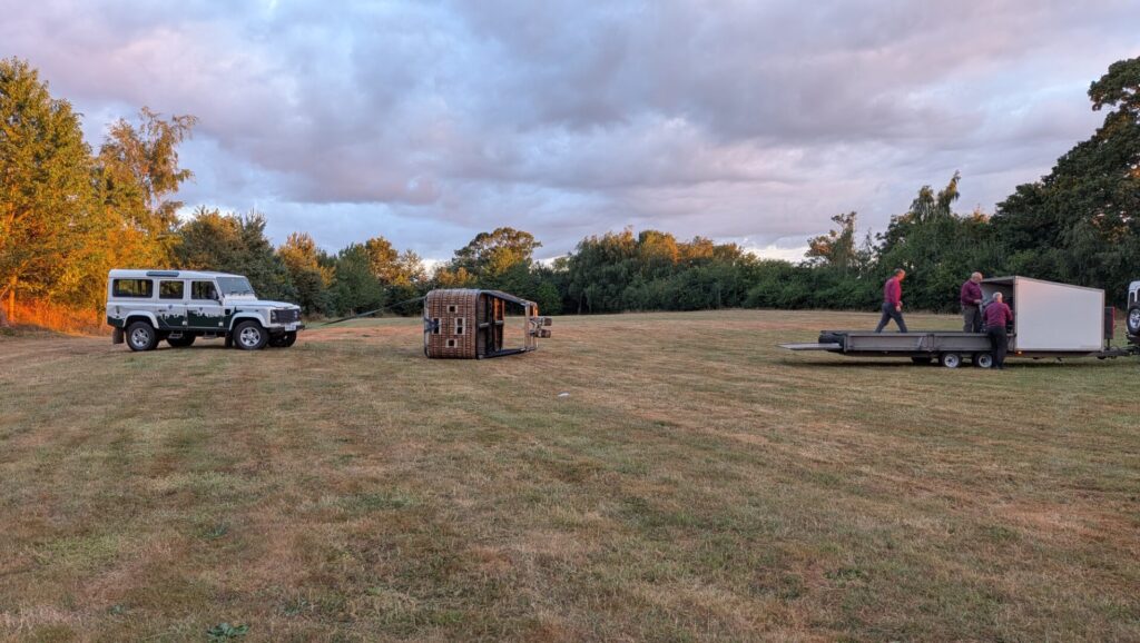 Photo of the hot air balloon basket on its side, tethered to an old style landrover. Three men are prepping the 'balloon' section for removal from a second landrover's trailer.